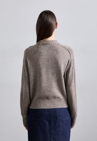 Nainen, jolla on pitkät suorat hiukset, pukeutuneena ruskeaan neulepaitaan ja tummansinisestä farkkuhameeseen, kuvattuna takaa yksinkertaista taustaa vasten.