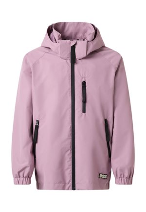 Jeune fille aux longs cheveux bouclés portant une veste à capuche mauve, des leggings noirs et des baskets blanches avec des semelles bleues, debout les mains dans les poches.