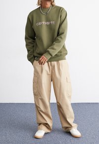 Oliivinvihreä collegepaita, jossa on violetinvärinen brändilogo, yhdistettynä beigeihin cargo-housuihin, joissa on suuret taskut, ja valkoisiin tennareihin, joissa on kumipohjat.