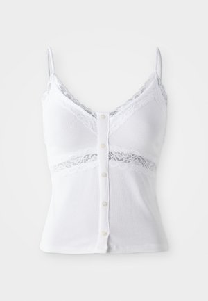 Bijela majica bez rukava s čipkastim detaljima i trakicama. Ima rebrastu teksturu i dekorativne dugmad duž prednje strane.