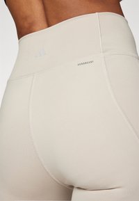 Beige high-waisted leggings with smooth texture. Features 'AEROREADY' branding and subtle seams for added design detail.