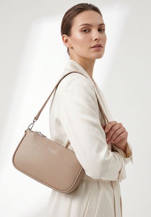 Femme en blazer crème portant un sac à main beige Diana & Co sur l'épaule devant un fond clair uni.