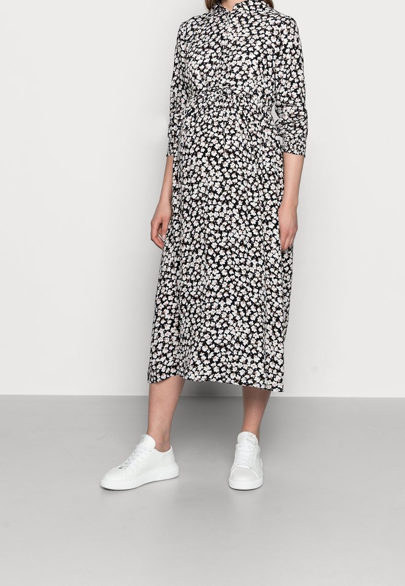 Robe florale noire avec des fleurs blanches et roses, dotée d'un col, de manches longues et d'une taille froncée, associée à des baskets blanches.