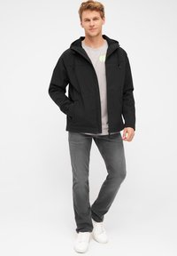 Giacca softshell nera con cappuccio, dotata di una cerniera frontale e tasche laterali, indossata su una maglietta grigia e jeans grigi, con sneaker bianche.
