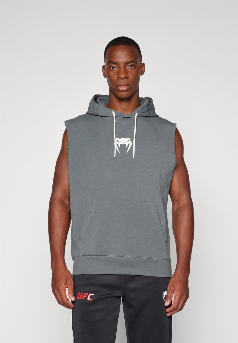Bezrukávová šedá mikina z mäkkej tkaniny, s predným vreckom a bielymi šnúrkami. Logo na hrudi, nosená s čiernymi nohavicami so značkou "UFC".