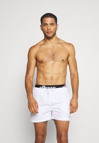 Weiße Badeshorts mit einem schwarzen elastischen Bund, der das "ellesse"-Logo zeigt. Leichter Stoff, Kordelzugverschluss und kurzer Schnitt.