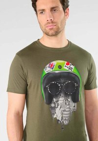 Camiseta verde oliva con un diseño gráfico de un casco con gafas de sol y detalles de un paisaje urbano, combinando color y elementos urbanos.