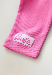 Lyserød ribbet stof med en hvid etiket, der har ordet "Barbie" skrevet i lyserød kursiv. Teksturen ser blød og strækbar ud.