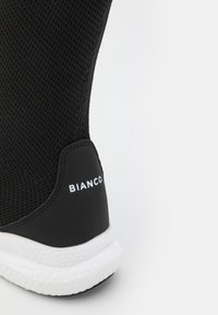 Zapatilla tipo calcetín negra con parte superior de malla texturizada, entresuela blanca a contraste y detalle de marca en el talón. Presenta una forma aerodinámica y deportiva.