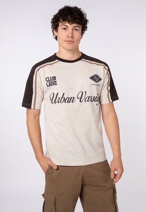 Young man with curly hair wearing a beige and black short-sleeve t-shirt with "Urban Varsity" text and brown cargo shorts, hands in pockets.