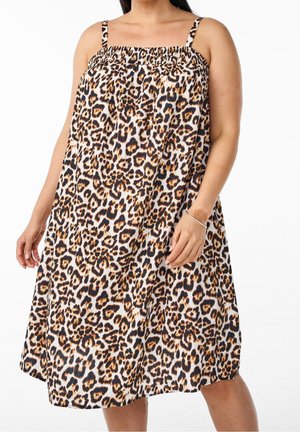 Kobieta ubrana w bezrękawową, sięgającą kolan sukienkę w wzór panterki, z cienkimi ramiączkami, stojąca na białym tle.