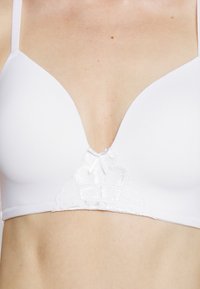 Sutiã acolchoado branco com um design em V, apresentando um delicado acabamento em renda e um pequeno laço ao centro. Textura de tecido suave.