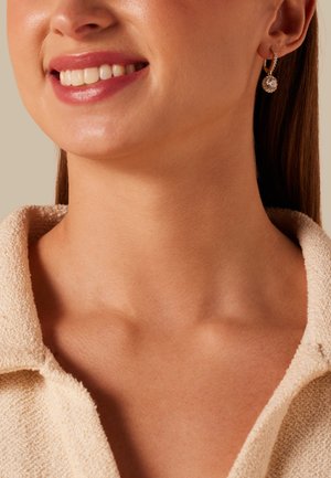 Lächelnde Frau mit strukturiertem beigen Oberteil mit Kragen und goldenen Creolen mit klaren runden Steinen vor neutralem Hintergrund.