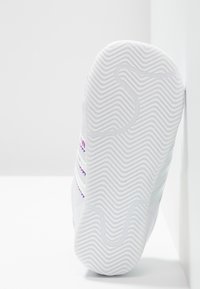 Vit sneakersula med zigzag-mönster, mot en vit bakgrund och vägg, som visar sidostänger i pastellgradientfärger.