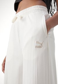 Detailní záběr osoby v bílých žebrovaných teplákách Puma s elastickým pasem a bočními kapsami, s jednou rukou v kapse.