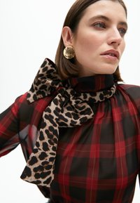 Červeno-čierna kockovaná blúzka s leopardím vzorom na mašličke, vyrobená z priesvitného materiálu a doplnená okrúhlymi zlatými náušnicami.