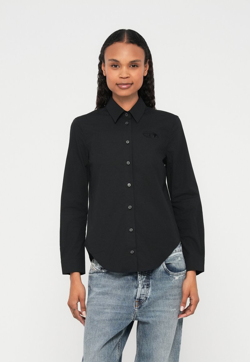 Melna garām piedurknēm krekls ar pogu priekšpusē, smailu apkakli un mazu logotipa detaļu. Kombinēts ar gaišas krāsas džinsiem.