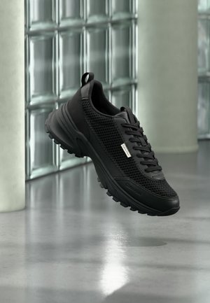 Schwarzer sportlicher Sneaker mit Mesh-Seitenteilen und dicker Sohle, der drinnen in der Nähe von Glasbausteinen und Säulen auf einem glatten Boden schwebt.