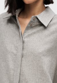 Grijs overhemd met knoopsluiting, gemaakt van een zachte, gestructureerde stof, voorzien van een kraag en twee knopen op de knoopsluiting. Het ontwerp is minimalistisch en op maat gemaakt.