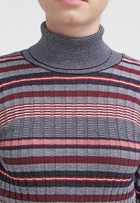 Virkat höghalstrad tröja med randiga mönster i röd, marinblå och vit. Ribbade texturer med en figurnära design som framhäver en hög krage.