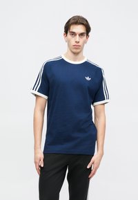 Námorníckomodrá tričko s krátkym rukávom a bielym lemovaním, s tromi bielymi pruhmi na pleciach. Na hrudi sa nachádza malé biele logo.