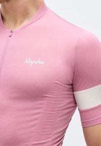 Κοντινή εικόνα ενός ατόμου που φοράει ροζ υφασμένο ποδηλατικό τζέρσεϊ με λευκή οριζόντια ρίγα στο μανίκι και το λογότυπο "Rapha" στο στήθος.