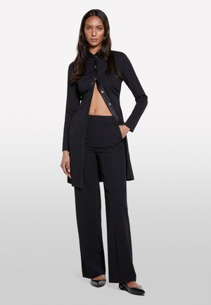 Camicia nera a maniche lunghe con colletto, dotata di bottoni dorati e un davanti aperto asimmetrico, abbinata a pantaloni neri larghi coordinati.