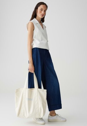 Cremfarbener Canvas-Shopper mit "BISOU" in Hellblau bedruckt, ausgestattet mit breiten Trägern und einem geräumigen Design, gehalten von einer Person in lässiger Kleidung.