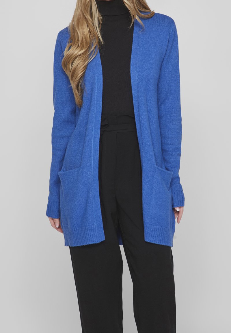 Blauer Strickcardigan mit offener Front, gerippten Bündchen, zwei vorderen Taschen und einer weichen Textur, kombiniert mit schwarzen Hosen.