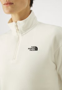 Maglione in pile con zip a quarti di crema, dal tocco morbido. Presenta un logo ricamato nero sul lato sinistro del petto e un alto colletto.