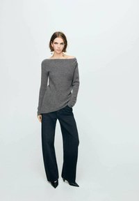 Suéter de punto gris sin hombros con un acabado texturizado, que presenta un dobladillo diagonal y detalles de botones, combinado con pantalones de pierna ancha oscuros y tacones.