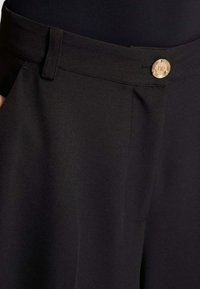 Pantaloni neri con una texture liscia, vita larga e un dettaglio con un bottone dorato con le iniziali "DR" sul davanti.