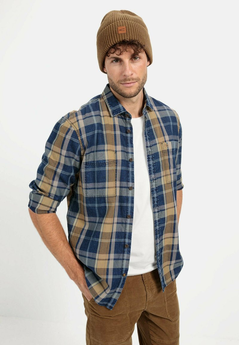 Gestrikte flanellen overhemd in blauw en beige met knoopsluiting en borstzak, gedragen over een witte t-shirt. Bruine gebreide beanie met logo.