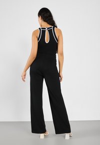 Combinaison noire à jambes larges, avec un contraste de finition blanche le long de l'encolure et un détail dos en trou de serrure. Tissu lisse et léger.