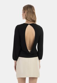 Sort langærmet bluse med en V-formet udsparing på ryggen, med rund halsudskæring og sammenkrøllet detalje ved taljen. Båret med en beige nederdel.