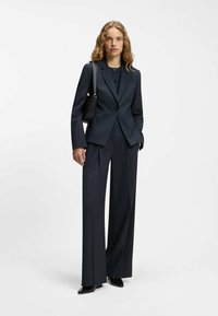 Abito sartoriale blu navy scuro composto da blazer aderente con un bottone singolo, pantaloni a gamba larga e tacchi neri lucidi. Completato da una piccola borsa nera.