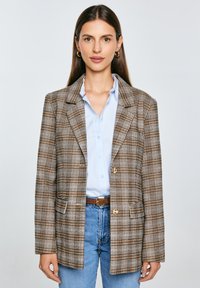 Hnědo-beige kostkovaný blazer s volným střihem, dvěma knoflíky a bočními kapsami, položený na světle modré košili a džínách.