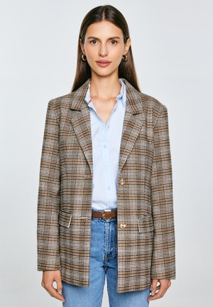 Blazer de cuadros marrón y beige con un corte amplio, dos botones y bolsillos laterales, combinado con una camisa azul clara y vaqueros.