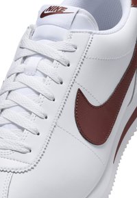 White Nike sneakers with a textured leather finish, featuring a maroon swoosh, flat laces, and a padded tongue for comfort.