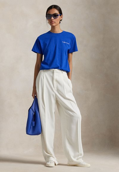 Sieviete, tērpusies zilā T-kreklā un baltās plašās biksēs, turot zilu somiņu, valkā saulesbrilles un baltas sporta kurpes.