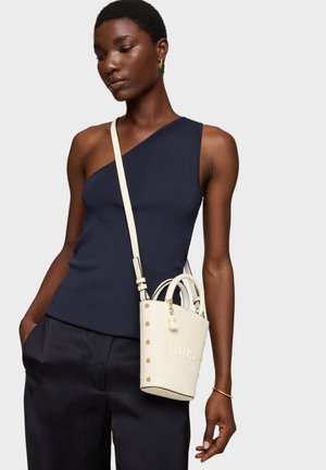 Donna che indossa un top monospalla blu navy e pantaloni neri, con una borsa a tracolla bianca con la scritta "TOUS" in rilievo sul davanti.