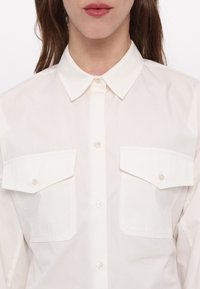 Femme portant une chemise blanche boutonnée avec un col pointu et deux grandes poches poitrine boutonnées, montrée du menton à la taille.