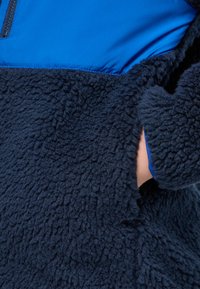 Veste en polaire bleu marine avec une couche extérieure texturée, comportant une section en tissu bleu lisse en haut et une poche latérale.