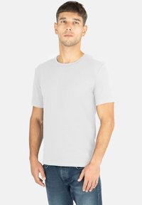 Rundhals, kurzärmliges weißes T-Shirt aus weicher Baumwolle mit einem dezenten Logodetail auf der Brust. Kombiniert mit dunkelblauen Jeans.