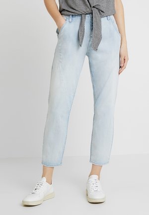 Pantalons en denim bleu clair avec une coupe décontractée, présentant des détails de couture en diagonale et deux poches latérales, associés à des baskets blanches.