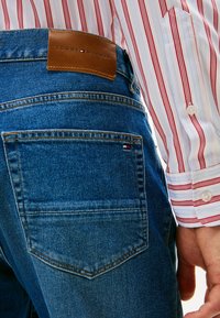 Vaqueros de mezclilla en azul medio con una etiqueta de cuero que lleva "TOMMY HILFIGER". El bolsillo trasero tiene un pequeño emblema de bandera. Camisa a rayas visible por el lado.