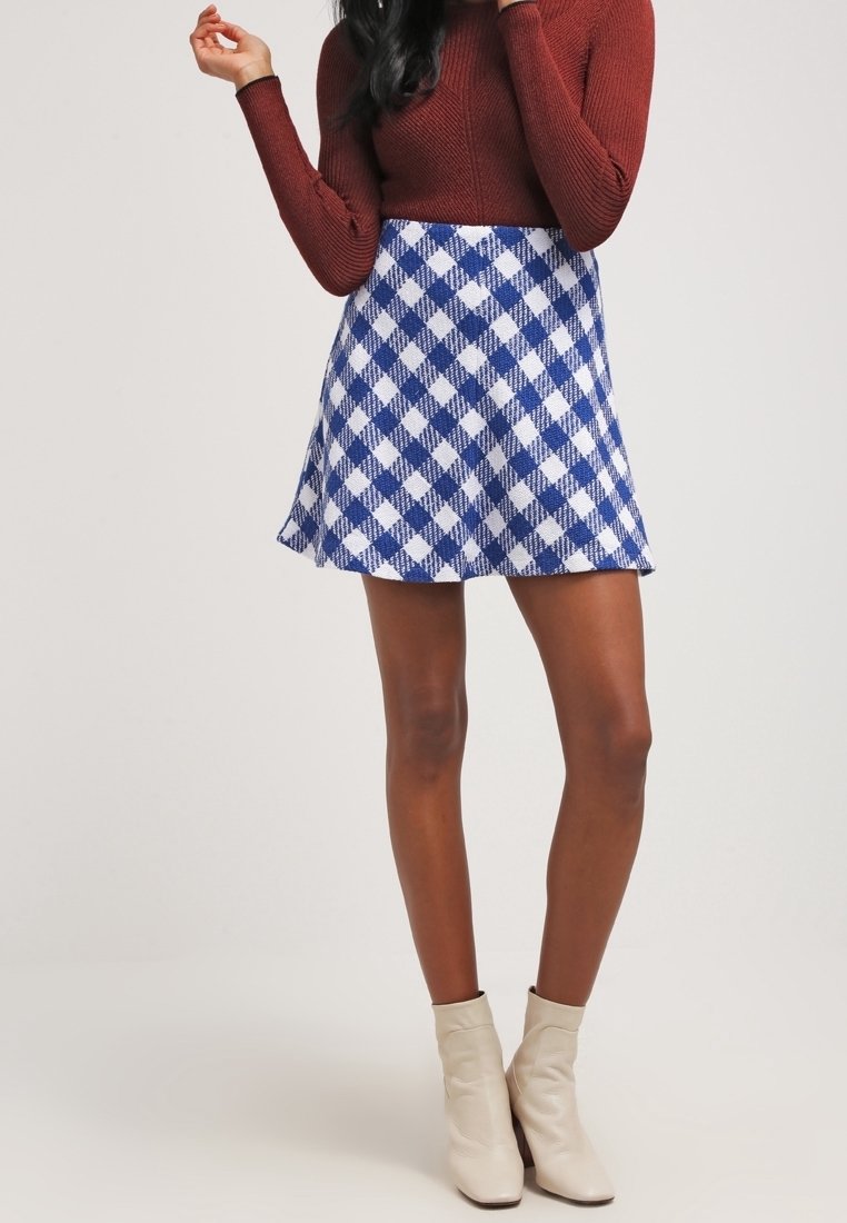 Femme portant une jupe à carreaux bleue et blanche, un haut marron à manches longues et des bottines beiges, debout avec une main levée.