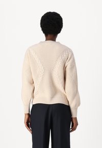 Pull à mailles crème avec un motif en câble texturé, épaules tombantes et ourlet côtelé. porté sur un pantalon sombre, montré de l'arrière.