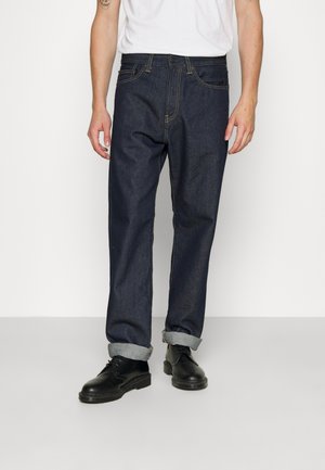 Personne portant un jean bleu foncé retroussé, des chaussures noires à lacets et une chemise blanche à manches courtes, debout devant un fond clair uni.