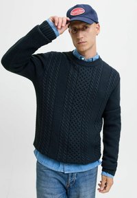 Pull bleu marine avec des motifs de tricot texturés et un ourlet côtelé, superposé sur une chemise en jean bleu clair, porté avec une casquette bleu marine.